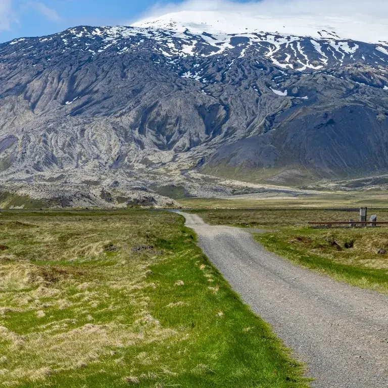 Schotterstraße führt durch grasige Ebenen auf das gewaltige, von Schnee bedeckte Snæfellsjökull-Massiv zu; klare Sicht auf gefurchte Lavaflanken – ruhige Anreise ins Hochland.
