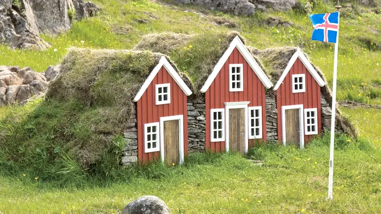 Drei rote, grasbedeckte Miniatur-Torfhäuser mit weißen Türen und Giebeln stehen auf einer Wiese; rechts weht eine kleine isländische Flagge. Die Szene vermittelt traditionelle Bauweise und ländliches Flair.