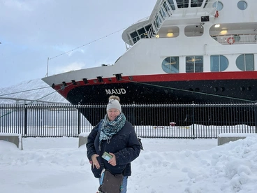 Eine Reisende steht warm eingepackt im Schnee vor einem Schiff im Hafen von Tromsø in Nordnorwegen. Das winterliche Licht und die verschneite Umgebung vermitteln das besondere Flair einer Schiffsreise über die Fjorde des Nordens – ein unvergessliches Erlebnis für Polar- und Norwegenliebhaber.