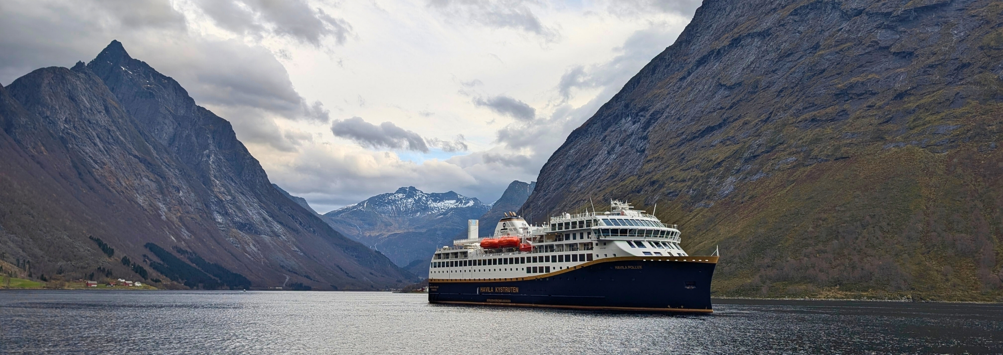Auf diesem Bild ist ein Schiff der „Havila Kystruten“ zu sehen, wie es durch die majestätische Landschaft des Hjørundfjords in Norwegen fährt. Das moderne Küstenschiff wird von den extrem steilen und imposanten Gipfeln der Sunnmøre-Alpen eingerahmt, was die spektakuläre Natur der norwegischen Fjorde verdeutlicht.