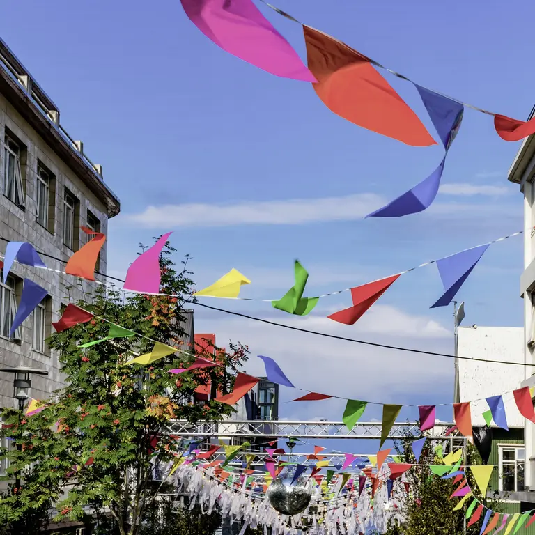 Bunte Wimpelketten spannen sich zwischen Häusern über eine Fußgängerzone in Reykjavík; sonniger Himmel, fröhliche Festival-Stimmung – Symbol für Kultur- und Stadtfeste in Island.