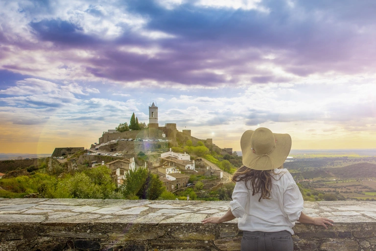 Blick auf das historische Dorf Monsaraz im Alentejo, Portugal, mit weiß getünchten Häusern und einer Burganlage auf einem Hügel. Eine Person mit Hut steht im Vordergrund und schaut über die Landschaft, während der Sonnenuntergang den Himmel in warme Farben taucht.
