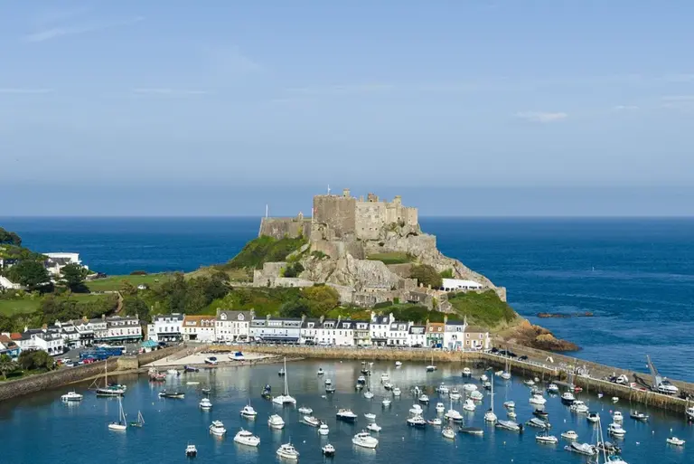 Panoramablick auf den Hafen von Jersey mit zahlreichen Segelbooten im ruhigen Wasser. Auf einer kleinen Halbinsel thront Elizabeth Castle über dem Meer, während sich weiße Häuser entlang der Küste unter blauem Himmel erstrecken.
