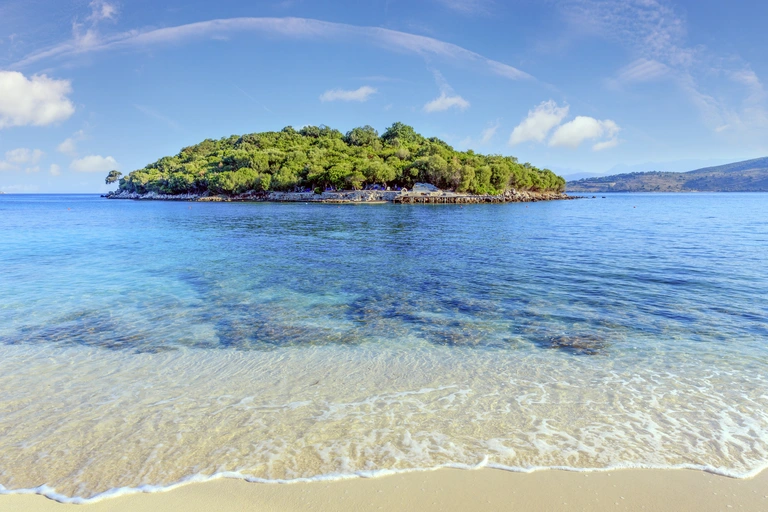 Paradiesische Badebucht mit türkisfarbenem Wasser und grüner Insel Eine kleine, dicht bewaldete Insel liegt in einem klaren, türkisblauen Meer. Im Vordergrund trifft sanftes Wellenrauschen auf einen hellen Sandstrand, darüber ein sonniger Himmel mit ein paar weißen Wolken.