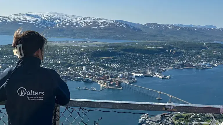 Panoramablick auf Tromsø Auf diesem Bild ist eine Frau zu sehen, wie sie von einem hohen Aussichtspunkt den Panoramablick auf die norwegische Stadt Tromsø genießt. Man erkennt die Stadt, die sich über eine Insel erstreckt, die markante Tromsø-Brücke und die umliegende Fjordlandschaft mit schneebedeckten Bergen im Hintergrund.