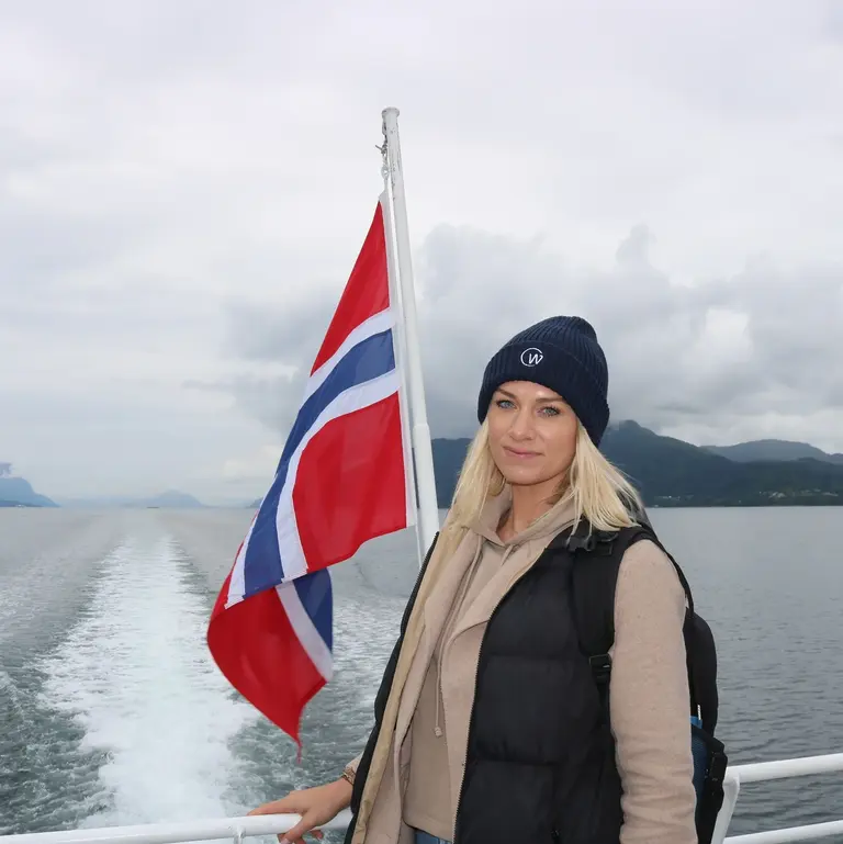 Fährfahrt in Norwegen Auf diesem Bild ist eine Frau zu sehen, die an Deck eines Schiffes vor einer wehenden norwegischen Flagge posiert. Im Hintergrund sieht man das Kielwasser des Schiffes und die in Dunst getauchte Fjordlandschaft, was die typische Atmosphäre einer Fährfahrt in Norwegen einfängt.