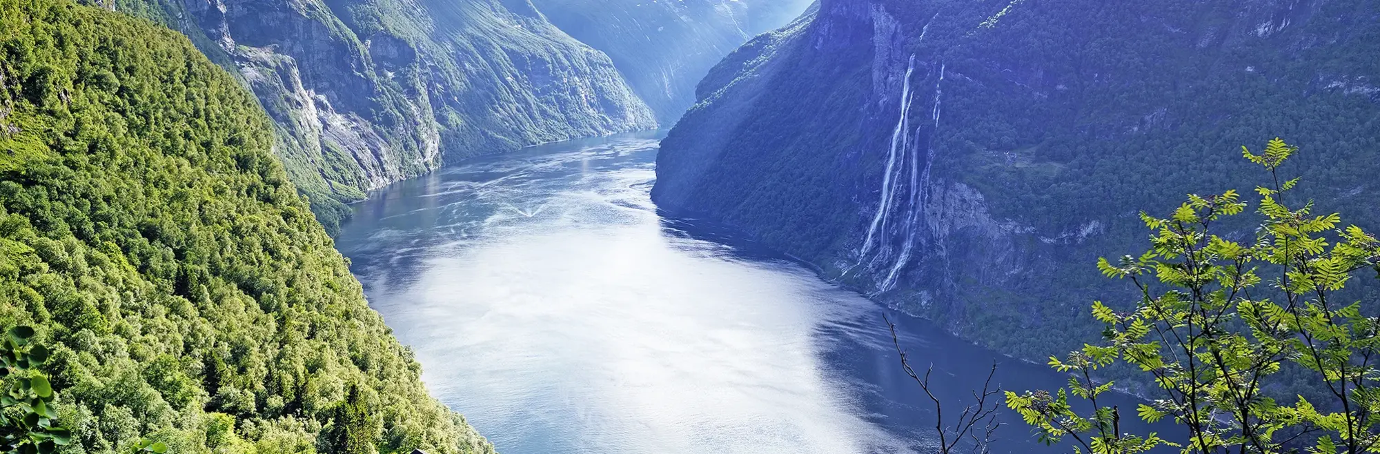 Geirangerfjord zwischen den Bergen - UNESCO-Weltnaturerbe mit Wasserfällen Das Bild zeigt den berühmten Geirangerfjord in Norwegen, umgeben von steilen, grünen Berghängen. Im Hintergrund stürzt der Wasserfall "Sieben Schwester" ins Tal. Diese spektakuläre Fjordlandschaft gehört zum UNESCO-Weltnaturerbe und ist ein Highlight jeder Norwegen-Rundreise.