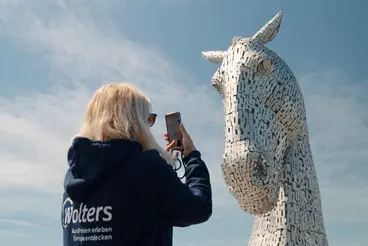 Die Kelpies in Schottland: Ein Besuch bei den majestätischen Pferdeskulpturen Eine Frau macht während ihrer Reise nach Schottland ein Foto von der beeindruckenden, aus Stahl gefertigten Pferdekopf-Skulptur der Kelpies in Falkirk, eine weltberühmte kulturelle Sehenswürdigkeit und ein Highlight für jeden Urlaub.