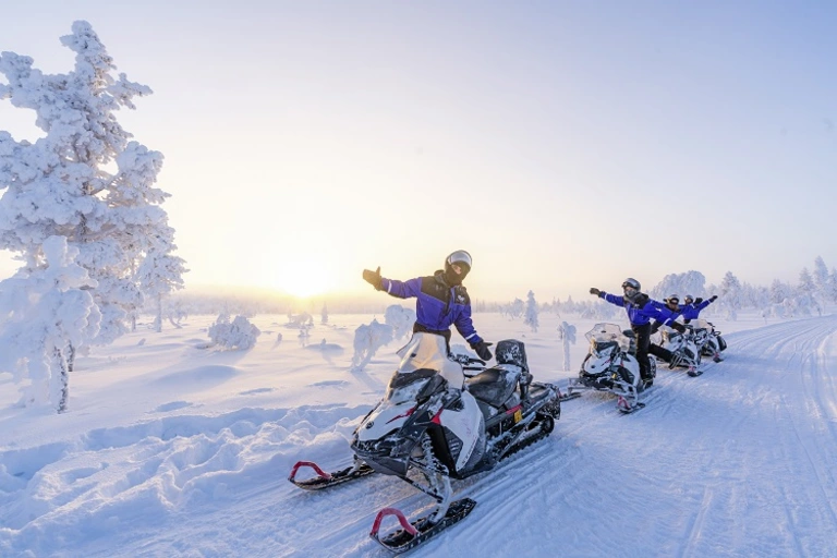 Schneemobil-Abenteuer in Lapplands Winterlandschaft Mehrere Personen fahren auf Schneemobilen durch eine verschneite, sonnenbeschienene Winterlandschaft, umgeben von frostigen Bäumen und glitzerndem Schnee.