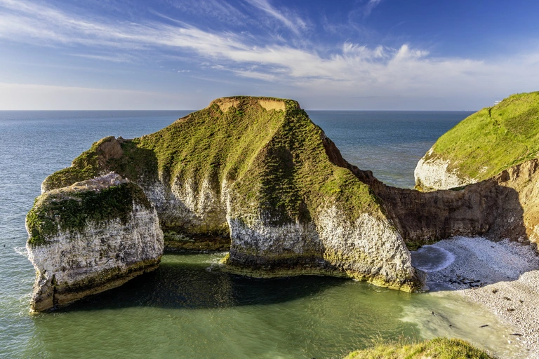 Steile, grün bewachsene Klippen an der Yorkshire Coast in Nordengland ragen über das Meer. Eine geschwungene Bucht mit hellem Sandstrand liegt zwischen den Felsen, während das klare Wasser und der weite Horizont die natürliche Küstenlandschaft Europas zeigen.