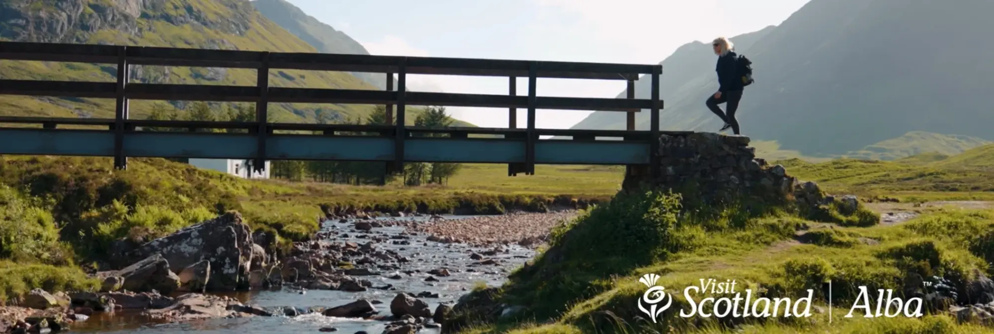 Eine blonde Frau überquert eine Holzbrücke über einen klaren Gebirgsbach inmitten der schottischen Highlands. Grüne Hügel und Bergketten bilden eine beeindruckende Kulisse unter blauem Himmel.