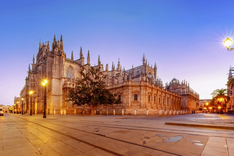 Kathedrale von Sevilla Sevillas Kathedrale bei Dämmerung, mit beleuchteter Fassade, gotischen Türmen und leerem Platz im Vordergrund
