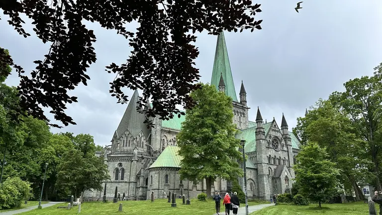 Der Nidarosdom in Trondheim Auf diesem Bild ist der beeindruckende Nidarosdom in Trondheim zu sehen, umgeben von einem grünen Park mit altem Baumbestand. Die Aufnahme an einem bewölkten Tag zeigt die majestätische gotische Architektur der Kathedrale mit ihren zahlreichen Türmen und den charakteristischen grünen Kupferdächern.