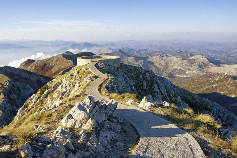 Atemberaubende Aussicht am Lovćen-Nationalpark in Montenegro Ein gewundener Steinweg führt über einen felsigen Bergkamm im Lovćen-Nationalpark in Montenegro. Am Ende des Weges liegt eine Aussichtsplattform mit weitem Blick über die umliegenden Berge und Täler unter klarem Himmel.