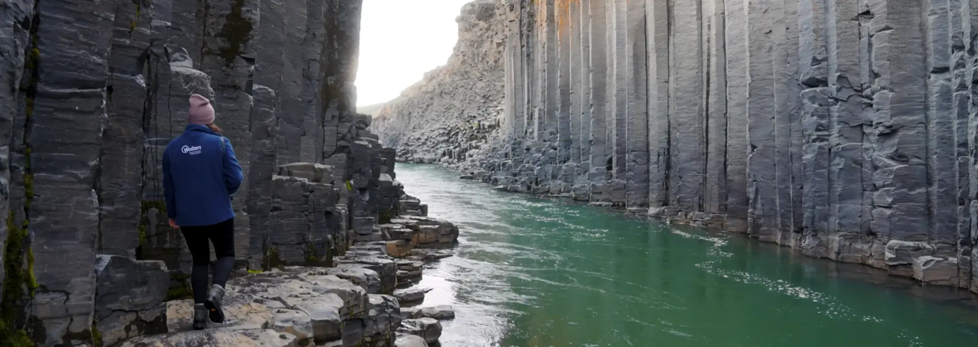 Wanderung im Basalt-Canyon Umgeben von beeindruckenden, sechseckigen Basaltsäulen wandert eine Frau durch den Stuðlagil Canyon in Island. Die Aufnahme fängt die einzigartige Schönheit dieser geologischen Formation ein, bei der der türkisfarbene Fluss einen eindrucksvollen Kontrast zu den dunklen, ebenmäßigen Steinsäulen bildet.