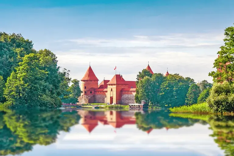 Trakai-Wasserburg: Historisches Juwel im Seenland Litauens Die mittelalterliche Wasserburg von Trakai steht auf einer Insel inmitten eines klaren Sees in Litauen. Das rotgedeckte Gemäuer spiegelt sich im ruhigen Wasser, eingerahmt von dicht bewachsenen grünen Bäumen unter einem hellen Sommerhimmel.