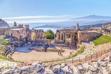 Das antike griechisch-römische Theater von Taormina auf Sizilien mit halbkreisförmigen Sitzreihen aus Stein. Im Hintergrund sind das tiefblaue Meer und der Ätna zu sehen, während die historische Ruine unter klarem Himmel in mediterraner Landschaft liegt.