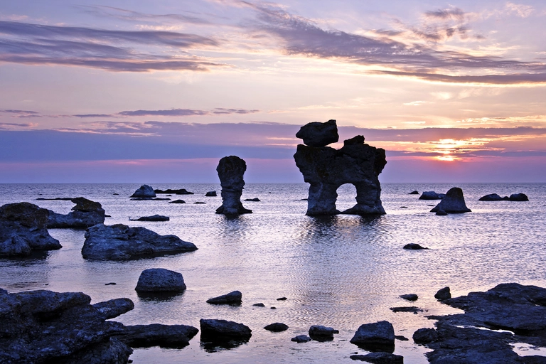 Markante Kalksteinfelsen, sogenannte Raukar, an der Küste von Gotland in Schweden bei Sonnenuntergang. Die Felsformationen ragen aus dem ruhigen Meer, während der Himmel in rosa und violetten Farben leuchtet und sich im Wasser spiegelt.