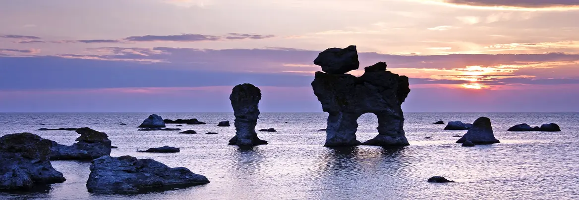 Im warmen Licht eines Sonnenuntergangs ragen eindrucksvolle Kalksteinfelsen aus dem Meer vor der Küste Gotlands in Schweden. Das Wasser schimmert in Violett- und Orangetönen, während die bizarren Felsformationen eine magische Stimmung erzeugen – ein beliebtes Reiseziel für Naturfreunde und Fotografen.