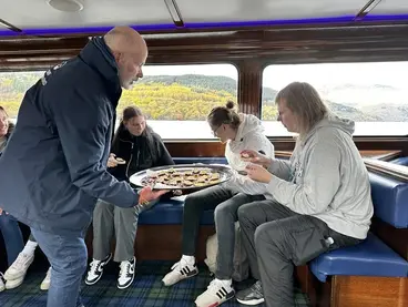 Gruppenreise in Schottland: Süße Pause bei einer Bootsfahrt in den Highlands Während einer unvergesslichen Bootsfahrt auf einem See in den schottischen Highlands genießt eine Reisegruppe eine süße, kulinarische Pause, während draußen die malerische Herbstlandschaft der Natur vorbeizieht und dieses gemeinsame Erlebnis zu einem Highlight im Urlaub macht.