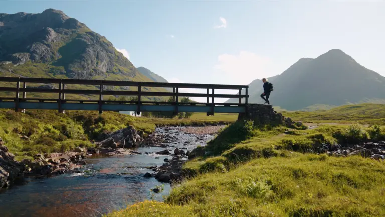 Eine Person überquert eine kleine Brücke über einen klaren Bach in den schottischen Highlands. Umgeben von sanften grünen Hügeln und markanten Bergsilhouetten zeigt die Szene eine ruhige, sonnige Landschaft, die typisch für die raue Schönheit Schottlands ist.