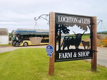 Schottisches Landleben: Ein Besuch der Lochton of Leys Farm & Shop Das Eingangsschild der "Lochton of Leys Farm & Shop" in Schottland wirbt mit der Silhouette eines Hochlandrinds für einen Besuch, während im Hintergrund ein Reisebus die Beliebtheit dieses Ausflugsziels für eine Reise in die schottische Kultur und das Landleben unterstreicht.