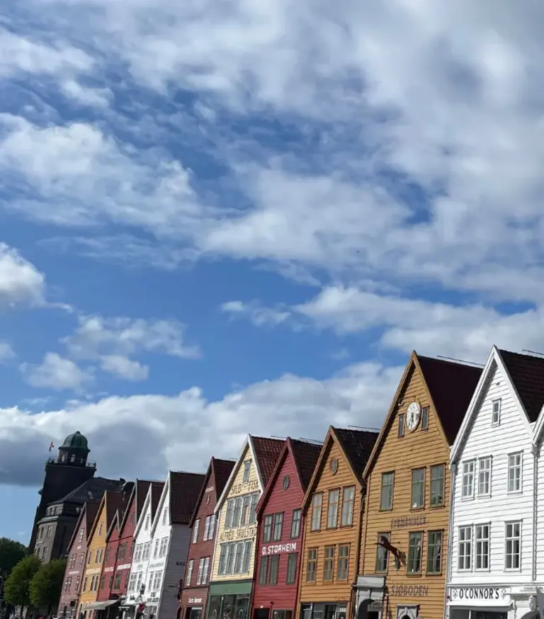 Die Giebelhäuser von Bryggen Auf dem Bild ist die berühmte, farbenfrohe Häuserzeile des Hanseviertels Bryggen in Bergen, Norwegen, unter einem malerischen Wolkenhimmel zu sehen. Die historischen Giebelhäuser aus Holz, die schief und eng aneinander stehen, repräsentieren ein UNESCO-Weltkulturerbe und die reiche Handelsgeschichte der Stadt.