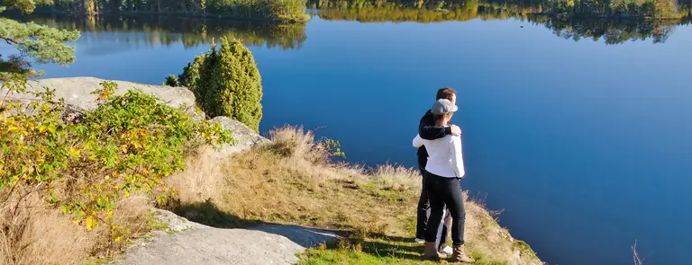 Zwei Personen umarmen sich auf einem Felsen mit Blick auf einen stillen See, umgeben von Bäumen und blauem Himmel - friedliche Momentaufnahme in der Natur Schwedens.