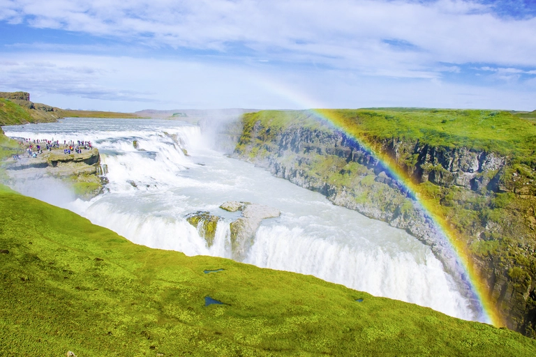 Der Gullfoss-Wasserfall in Island stürzt in zwei Stufen in eine tiefe Schlucht und erzeugt dichten Sprühnebel. Ein farbenfroher Regenbogen spannt sich über den Wasserfall, während Besucher auf einer Aussichtsterrasse die beeindruckende Naturkulisse umgeben von grünen Wiesen beobachten.