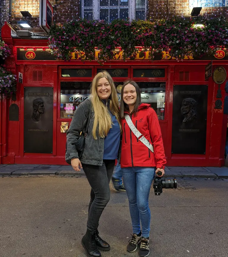 Vor der ikonischen roten Fassade des weltberühmten „The Temple Bar“ Pubs in Dublin posieren hier zwei Frauen für ein Erinnerungsfoto. Die nächtliche Beleuchtung und die fröhliche Stimmung der beiden fangen die lebendige Atmosphäre dieses beliebten touristischen Hotspots perfekt ein.