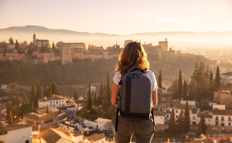 Eine Frau mit Rucksack blickt von einem Aussichtspunkt über Granada in Spanien. Vor ihr liegt die im Morgenlicht leuchtende Stadt mit der Alhambra auf einem Hügel, umgeben von Zypressen und sanften Dunstschichten. Die Szene vermittelt Ruhe, Fernweh und die historische Atmosphäre Andalusiens.