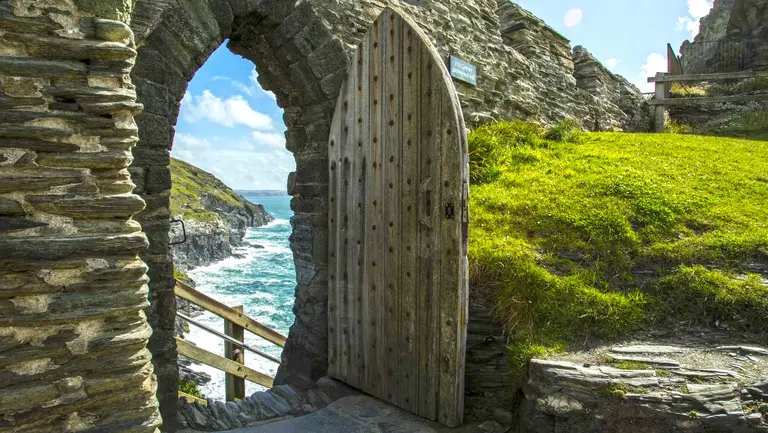 Blick durch einen alten Steintorbogen von Tintagel Castle auf die raue Atlantikküste mit Wellen und Felsen.