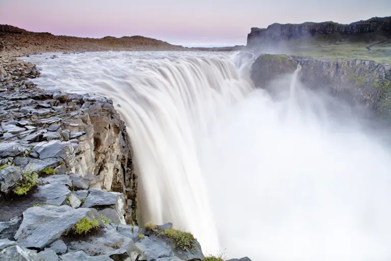 Der Gullfoss Wasserfall in Island stürzt in mehreren Stufen kraftvoll in eine tiefe Schlucht. Weißes Wasser fällt über breite Felskanten, umgeben von karger, vulkanischer Landschaft unter leicht bewölktem Himmel.