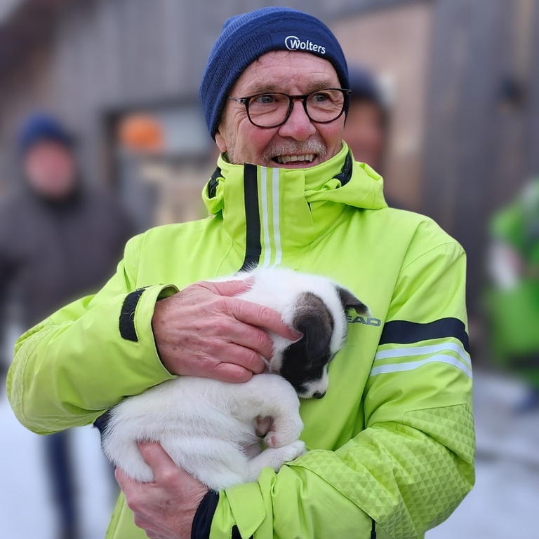 Auf diesem Bild ist ein Mann zu sehen, der in warmer Winterkleidung liebevoll einen kleinen, schlafenden Husky-Welpen in seinen Armen hält. Der Mann lächelt glücklich, was die herzliche und unvergessliche Begegnung mit dem Tier auf einer Farm in einer verschneiten Umgebung unterstreicht.
