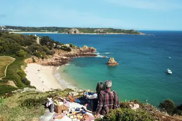 Eine Person sitzt auf einer Klippe auf Jersey und blickt auf eine geschützte Bucht mit Sandstrand und türkisblauem Meer. Im Vordergrund steht ein Picknickkorb mit Speisen, im Hintergrund erstrecken sich Felsen, Küstenvegetation und die britische Kanalinsel unter blauem Himmel.