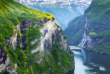 Blick auf den Geirangerfjord in Norwegen mit steilen, grün bewachsenen Felswänden, die direkt ins tiefblaue Fjordwasser abfallen. Mehrere schmale Wasserfälle stürzen von den Bergen ins Tal, während im Hintergrund schneebedeckte Gipfel zu sehen sind.