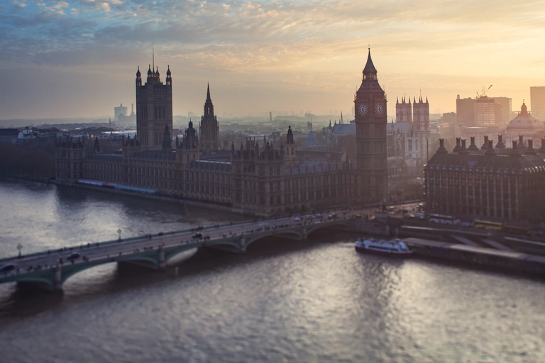 Panoramablick auf das Houses of Parliament und den Big Ben in London bei Sonnenaufgang. Die Westminster Bridge führt über die ruhig wirkende Themse, während warmes Morgenlicht die historischen Gebäude und die Skyline der Stadt stimmungsvoll beleuchtet.