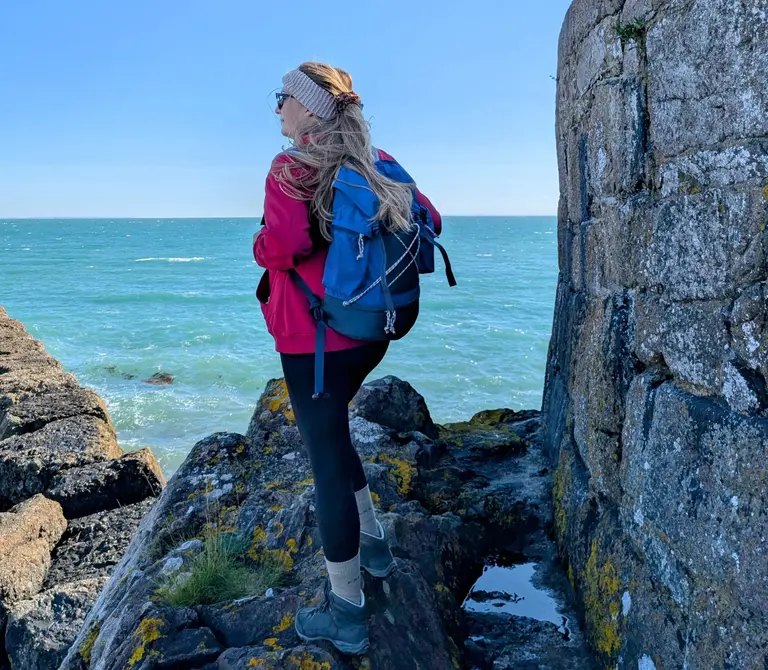 Wandern Jersey Eine Frau, die mit Wanderausrüstung auf einem Felsen auf Jersey steht und ihren Blick in Richtung des blauen Ozeans gewandt hat