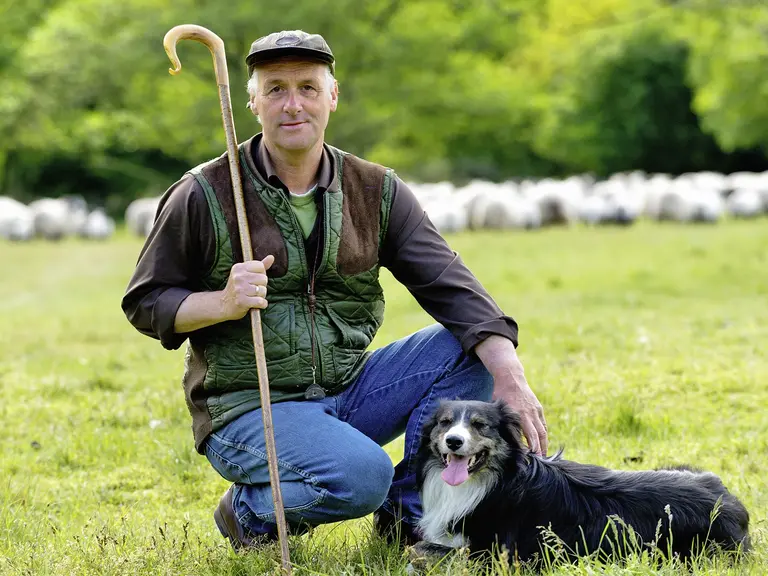 Ein schottischer Schäfer kniet auf einer Wiese neben seinem Hütehund, im Hintergrund weiden Schafe. Die Szene zeigt das traditionelle Landleben inmitten