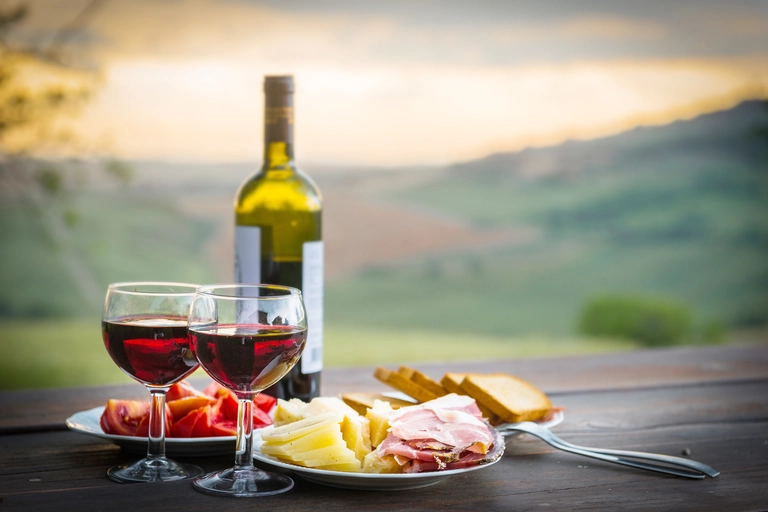 Zwei Gläser Rotwein mit Käse, Schinken, Brot und Paprika, serviert auf einem Holztisch in der toskanischen Landschaft.