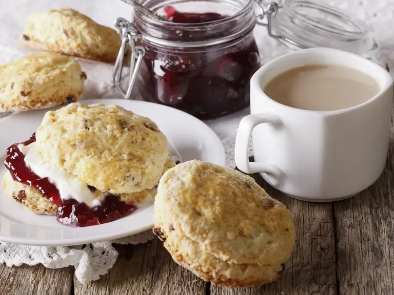 Teller mit frisch gebackenen Scones, bestrichen mit Clotted Cream und Erdbeermarmelade, daneben eine Tasse Tee. Typisches britisches Gebäck für den Nachmittagstee in Schottland.