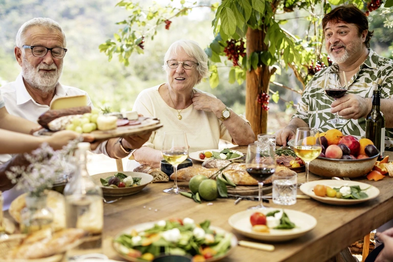 Ältere Menschen genießen gemeinsam ein italienisches Essen im Freien mit Wein, Brot, Obst und Salat – geselliges Beisammensein in mediterraner Atmosphäre.