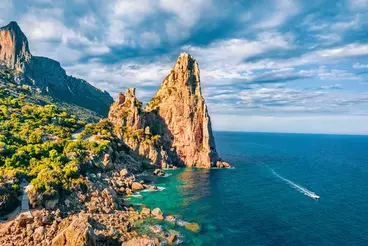 Steile Felsformationen an der Küste von Sardinien ragen über das türkisfarbene Mittelmeer. Zwischen grüner Macchia und schroffen Klippen verläuft die Küstenlinie unter blauem Himmel, während ein Boot eine Spur im Wasser hinterlässt.