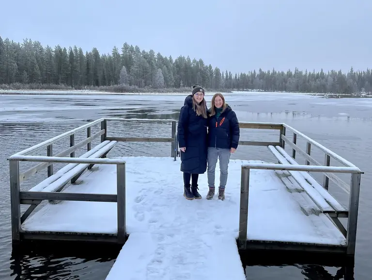 Winterzauber in Lappland: Ein unvergesslicher Moment am gefrorenen See Zwei Frauen genießen während ihrer Winter-Reise nach Lappland, Finnland, die stille Natur und stehen gemeinsam auf einem schneebedeckten Holzsteg, der in einen teilweise zugefrorenen See ragt, was dieses Abenteuer in der arktischen Landschaft zu einem unvergesslichen Urlaub macht.