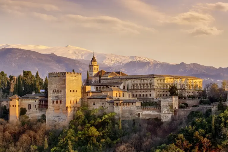 Die Alhambra in Granada, Andalusien, erhebt sich majestätisch über der Stadt. Die historische Palast- und Festungsanlage aus maurischer Zeit liegt eingebettet in grüne Hügel, mit der Sierra Nevada im Hintergrund unter warmem Abendlicht.