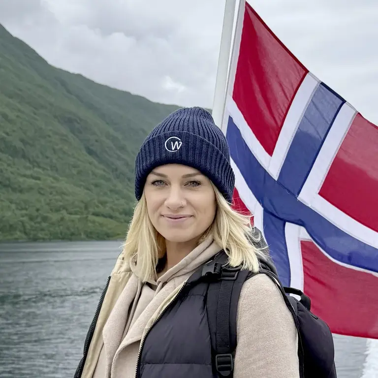 Unterwegs in Norwegen - Küstenreise mit Havila erleben Eine Reisende steht auf einem Schiff vor einer norwegischen Flagge und blickt auf die grüne Fjordlandschaft. Das Foto symbolisiert Abenteuerlust und nordische Gelassenheit - ideal für alle, die Norwegen per Postschiff entdecken möchten.