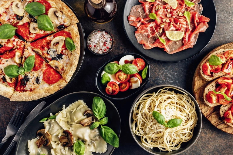 Auswahl typischer italienischer Gerichte auf einem Tisch angerichtet: Pizza mit Tomaten und Basilikum, Pasta mit Pesto, Ravioli, Antipasti und frische Zutaten wie Chili und Kräuter. Die mediterrane Küche Italiens steht für Genuss, Vielfalt und traditionelle Spezialitäten.