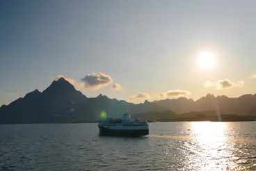 Mit dem Schiff durch Norwegen – Abendstimmung vor den Lofoten Ein Küstenschiff fährt bei tief stehender Sonne über das ruhige Meer vor den Lofoten in Norwegen. Die Silhouetten der markanten Berge zeichnen sich dunkel gegen den goldenen Himmel ab, während sich das Sonnenlicht glitzernd auf der Wasseroberfläche spiegelt.