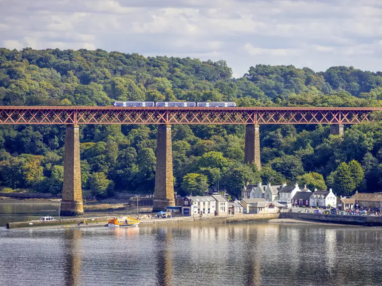 Ein Zug fährt über die hohe Forth Bridge bei South Queensferry, unter der sich das ruhige Wasser des Firth of Forth erstreckt. Im Vordergrund liegen kleine Häuser und Boote am Ufer