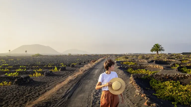 Person in vulkanischer Landschaft Eine Person steht auf einem Weg in einer weiten Landschaft mit dunklem, vulkanischen Boden und hält einen Strohhut in der Hand. Neben dem Weg befinden sich kleine Steinmauern, umgeben von Felden mit Pflanzen. In der Ferne sind Berge und eine Palme zu sehen.